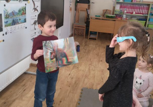 Widok na chłopca, który trzyma w rękach obrazek dinozaura w 3D i dziewczynkę w okularach. W tle tablica tematyczna o dinozaurach.